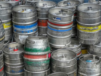 A collection of metal beer kegs stacked together, each with colored stripes of yellow, blue, red, and green marking lines around them. The kegs have a cylindrical shape and are made of stainless steel, showing signs of wear and tear with various engravings and labels visible on their surfaces.