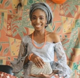 A smiling individual wearing traditional clothing, including a headwrap and an off-shoulder dress accented with lace. The person is accessorized with orange beads and is holding a decorated clutch. The background features a colorful, abstract-patterned fabric with leaves and geometric shapes.