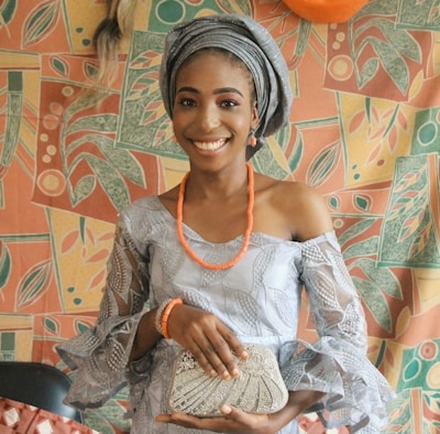 A smiling individual wearing traditional clothing, including a headwrap and an off-shoulder dress accented with lace. The person is accessorized with orange beads and is holding a decorated clutch. The background features a colorful, abstract-patterned fabric with leaves and geometric shapes.