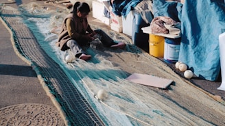A person working on repairing a large fishing net laid out on the ground, surrounded by various items such as buckets and containers. The setting appears to be outdoors in a sunny area, with shadows visible on the ground.