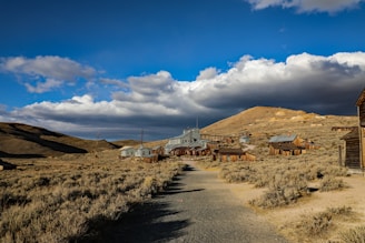 A deserted ghost town with wooden buildings and cabins arranged along a dirt path, set against a backdrop of arid hills and a dramatic sky filled with thick, dark, and fluffy clouds. The buildings are spaced out and the landscape is sparse with dry grass and shrubs.