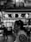 A black and white image featuring a person in the foreground with a blurred background of a bustling urban street. The background includes illuminated signs with Asian characters, suggesting a lively marketplace or city area. The person appears to be walking, with other silhouettes of people visible in the distance.