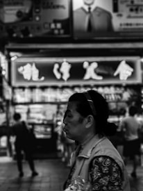 A black and white image featuring a person in the foreground with a blurred background of a bustling urban street. The background includes illuminated signs with Asian characters, suggesting a lively marketplace or city area. The person appears to be walking, with other silhouettes of people visible in the distance.