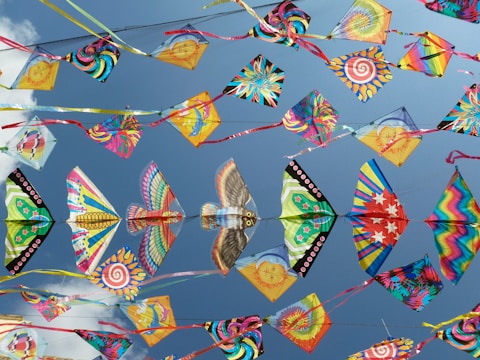 A collection of colorful kites with various patterns, including swirls, stars, and animal designs, flying against a bright blue sky.