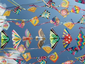 A collection of colorful kites with various patterns, including swirls, stars, and animal designs, flying against a bright blue sky.