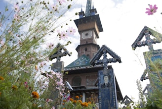 An intricately designed wooden church with a tall spire rises amidst colorful flowers and artistically decorated tombstones. The architecture features rich patterns and vibrant colors, typical of unique cultural heritage sites.