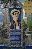 A vibrant, colorful gravestone featuring a carved portrait of a man wearing traditional clothing and a hat. The gravestone showcases intricate patterns and a poem or inscription written on it. It is situated in a cemetery with similarly styled graves in the background.