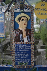 A vibrant, colorful gravestone featuring a carved portrait of a man wearing traditional clothing and a hat. The gravestone showcases intricate patterns and a poem or inscription written on it. It is situated in a cemetery with similarly styled graves in the background.
