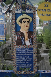 A vibrant, colorful gravestone featuring a carved portrait of a man wearing traditional clothing and a hat. The gravestone showcases intricate patterns and a poem or inscription written on it. It is situated in a cemetery with similarly styled graves in the background.