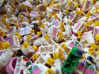 A large pile of LEGO mini-figure torsos featuring a design with a pink vest and black bow tie. The torsos have yellow hands, predominantly facing in different directions, and are scattered randomly, creating a chaotic and colorful scene.