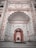 An intricately designed architectural entrance featuring ornate floral patterns and Arabic script. The archway is detailed with carved designs and has a series of smaller arches leading inward. The color scheme includes warm earth tones with a focus on symmetry and detailed craftsmanship.