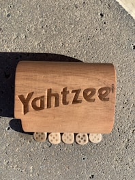 A wooden block with the word 'Yahtzee' engraved on it is lying on a rough, textured concrete surface. Below the block, there are five wooden pieces resembling dice, each showing five dots.