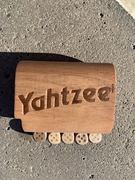 A wooden block with the word 'Yahtzee' engraved on it is lying on a rough, textured concrete surface. Below the block, there are five wooden pieces resembling dice, each showing five dots.