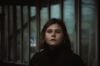 A person with short, brown hair and hoop earrings stands in front of a row of tall windows, appearing contemplative and serene. The background is slightly blurred, giving a soft focus on the person.