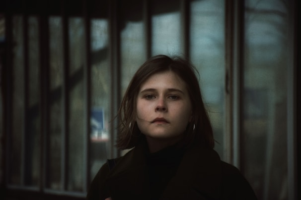 A person with short, brown hair and hoop earrings stands in front of a row of tall windows, appearing contemplative and serene. The background is slightly blurred, giving a soft focus on the person.