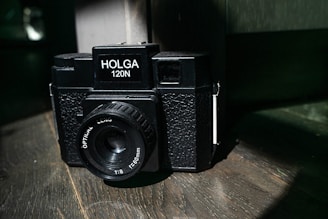 A vintage-style black Holga 120N camera with an optical lens sits on a wooden surface, partially illuminated by a beam of light. The camera's textured body and simple design evoke a classic, analog feel.