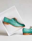 Elegant teal leather shoes with decorative perforations, set against a minimalist white backdrop. One shoe is elevated in a tilted white box, showcasing its polished wooden sole, while the other remains on the surface.