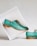 Elegant teal leather shoes with decorative perforations, set against a minimalist white backdrop. One shoe is elevated in a tilted white box, showcasing its polished wooden sole, while the other remains on the surface.