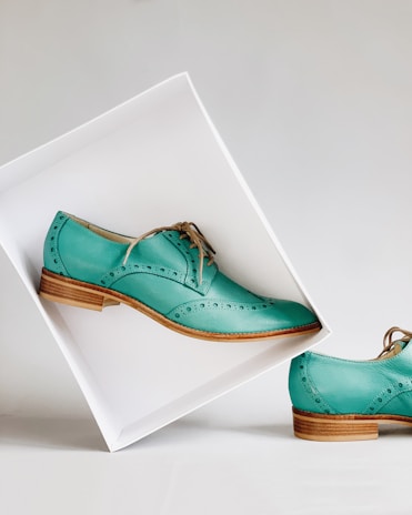 Elegant teal leather shoes with decorative perforations, set against a minimalist white backdrop. One shoe is elevated in a tilted white box, showcasing its polished wooden sole, while the other remains on the surface.