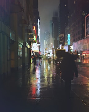 Wide shot of a rainy city street at night with glowing neon reflections on wet pavement.