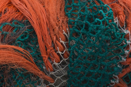 An intricate tangle of brightly colored fishing nets with orange and teal threads interwoven. The nets appear dense and textured, with strands and loops creating a complex pattern.