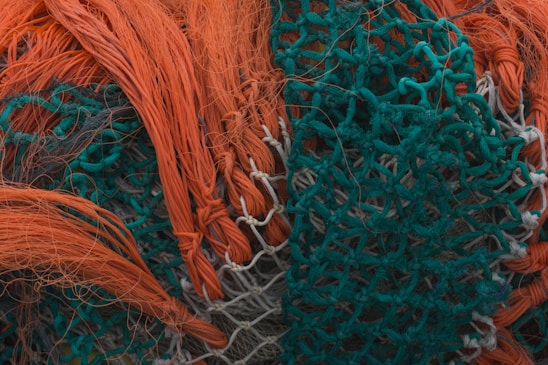 An intricate tangle of brightly colored fishing nets with orange and teal threads interwoven. The nets appear dense and textured, with strands and loops creating a complex pattern.
