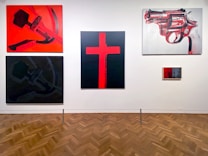 A gallery wall featuring five paintings with bold color contrasts. The upper left painting displays a stylized sickle and hammer on a vibrant red background. Below it is a darker, muted version of the same imagery. The center painting showcases a large red cross on a black background. On the upper right, a detailed image of a revolver is illustrated in shades of red and gray. A small, framed piece with red and black tones is also visible on the lower right.