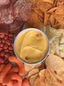 A central bowl of creamy, yellow cheese dip surrounded by an assortment of snacks including ridged potato chips, cheese curls, spicy puffed snacks, tortilla chips, and reddish seasoned chips.