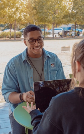 Two people are sitting at an outdoor table, with one person facing the camera and the other with their back to it. The person facing the camera is wearing glasses and a denim shirt, and appears to be smiling and engaged in conversation. They are sitting in a park with trees and parked cars visible in the background. A laptop is on the table between them.