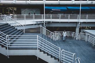A multi-level architectural space with a series of steps and railings, featuring a man walking along the pathway. The area has modern design elements and is situated under an overpass with businesses visible in the background.