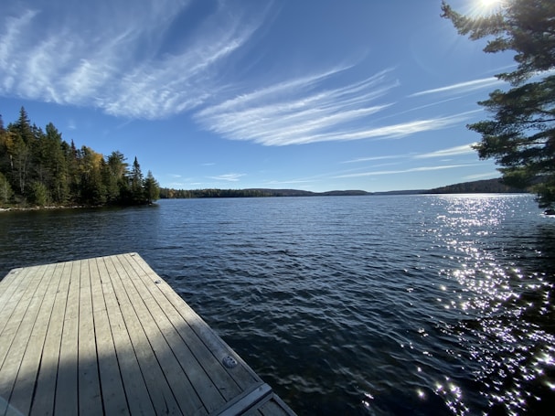 A serene lake extends towards the horizon, bordered by lush green forests on either side. The sky is bright blue with long, wispy clouds, and the sunlight sparkles off the water's surface. A wooden dock juts out into the lake, inviting tranquility and reflection, while the trees frame the scene beautifully.