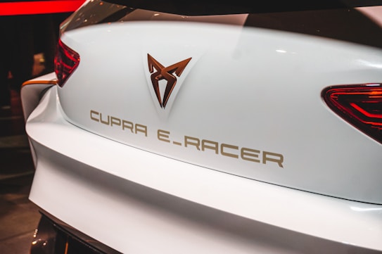 The image depicts the rear end of a white car displaying the logo and text 'CUPRA E-RACER'. The vehicle features a modern design with sleek brake lights positioned at the sides.