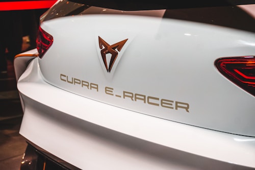 The image depicts the rear end of a white car displaying the logo and text 'CUPRA E-RACER'. The vehicle features a modern design with sleek brake lights positioned at the sides.