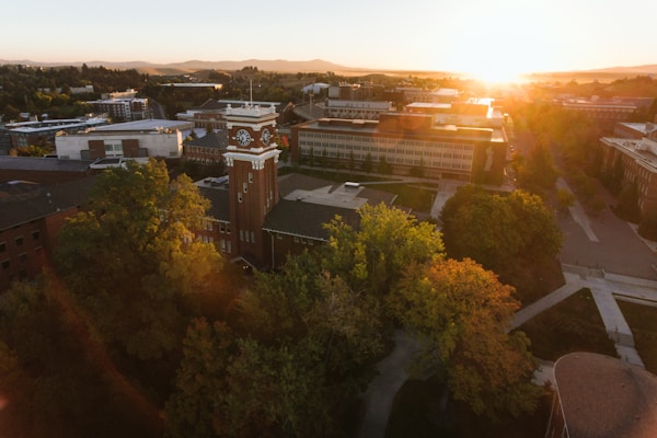 Aerial view of a university campus at sunset, featuring a large clock tower surrounded by multiple academic buildings and lush greenery. Sunlight bathes the scene with a warm glow while casting long shadows across walkways and structures.