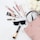 A selection of makeup products including lip gloss, mascara, eyeliner, foundation, and a makeup brush. These items are arranged neatly around a pair of stylish black sunglasses and a blush pink handbag on a white surface. In the top right corner, there is a bouquet of white roses.