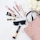 A selection of makeup products including lip gloss, mascara, eyeliner, foundation, and a makeup brush. These items are arranged neatly around a pair of stylish black sunglasses and a blush pink handbag on a white surface. In the top right corner, there is a bouquet of white roses.