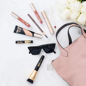 A selection of makeup products including lip gloss, mascara, eyeliner, foundation, and a makeup brush. These items are arranged neatly around a pair of stylish black sunglasses and a blush pink handbag on a white surface. In the top right corner, there is a bouquet of white roses.