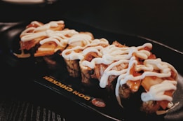A plate with pieces of sushi topped with a generous amount of white sauce, on a dark serving dish with a restaurant logo partially visible.