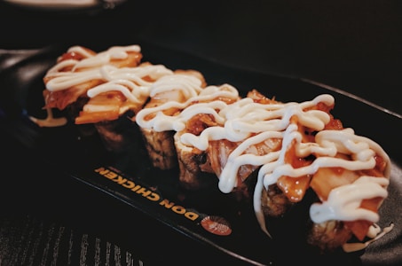 A plate with pieces of sushi topped with a generous amount of white sauce, on a dark serving dish with a restaurant logo partially visible.