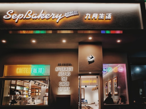 A cozy bakery shopfront named SepBakery is illuminated with warm lighting and colorful neon signs. The entrance features bright signs that say 'COFFEE' and 'AFTERTASTE JUST AS THE SAVOR OF LIFE.' Inside, the counter area is visible with shelves stocked with products, and there are customers seated inside enjoying their drinks.