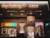 A cozy bakery shopfront named SepBakery is illuminated with warm lighting and colorful neon signs. The entrance features bright signs that say 'COFFEE' and 'AFTERTASTE JUST AS THE SAVOR OF LIFE.' Inside, the counter area is visible with shelves stocked with products, and there are customers seated inside enjoying their drinks.