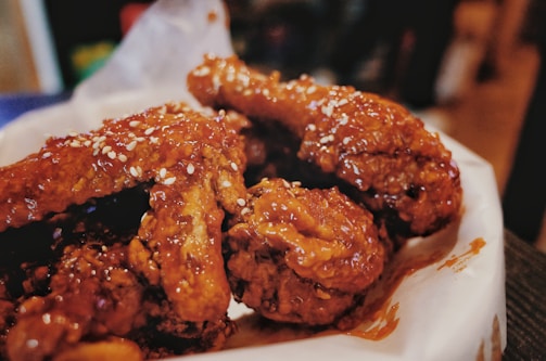Crispy, golden-brown fried chicken pieces are covered with a glossy, slightly sticky sauce, sprinkled with sesame seeds, and placed on a paper-lined tray.