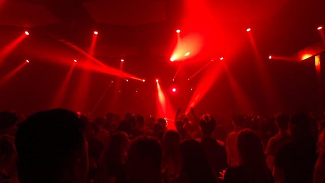A crowd of people is gathered in a dimly lit room with vibrant red lighting and multiple beams of light projecting onto the audience. The atmosphere suggests a lively event, possibly a concert or nightclub, with silhouettes of individuals enjoying the music and ambiance.