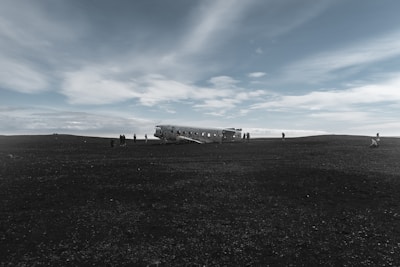 A desolate landscape featuring a crashed airplane fuselage rests on a barren dirt plain under a wide sky filled with soft, wispy clouds. Several people are scattered around the wreckage, some approaching the plane and others standing at a distance, while the expansive sky creates a sense of openness.