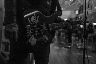 A person is holding an electric bass guitar with skull and other stickers on it, standing in what seems to be a dimly lit venue. The focus is on the guitar and the person's hand, with blurred figures and lights in the background creating a moody atmosphere.