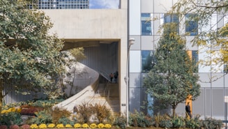 A modern architectural structure features a concrete ramp alongside a facade composed of glass panels. The landscape is enhanced by various trees and a well-maintained garden with colorful flowers. A person is walking down the ramp, and another is near the building, adding life to the urban scene. The composition balances natural elements with urban architecture.