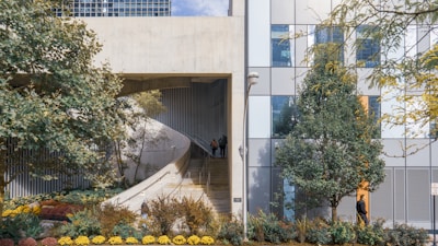 A modern architectural structure features a concrete ramp alongside a facade composed of glass panels. The landscape is enhanced by various trees and a well-maintained garden with colorful flowers. A person is walking down the ramp, and another is near the building, adding life to the urban scene. The composition balances natural elements with urban architecture.