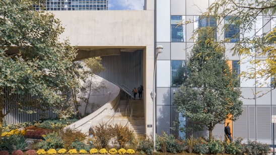 A modern architectural structure features a concrete ramp alongside a facade composed of glass panels. The landscape is enhanced by various trees and a well-maintained garden with colorful flowers. A person is walking down the ramp, and another is near the building, adding life to the urban scene. The composition balances natural elements with urban architecture.