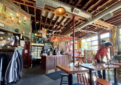 A cozy cafe with a rustic and eclectic interior, featuring wooden beams and exposed ductwork on the ceiling. The cafe has a long counter with shelves filled with beverages and two staff members working behind it. Tables and chairs are scattered throughout, and a person is seen setting a table. The decor includes colorful posters, flags, and chandeliers with warm lighting. Large windows allow natural light to fill the space.