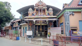 A Hindu temple with intricate carvings and colorful statues is set in an urban environment. The entrance features traditional architectural elements with brightly painted figures of deities. A canopy extends over the gate, and informative signs are displayed near the entrance. The surroundings include red metal barricades and a large tree to the side.